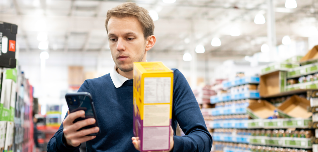 Man grocery shopping comparing food label with prices on his phone – representing the uncommitted consumer.