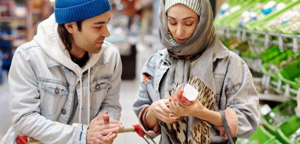 Two shoppers in a grocery store aisle review a product label together, with a cart full of groceries beside them, highlighting mindful purchasing and product transparency.